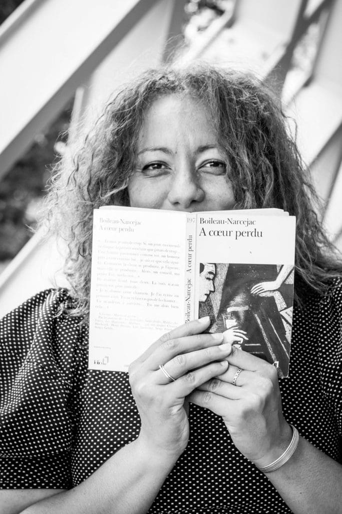A black and white image of a woman holding open a French novel in front of her with both hands. She is peering out over the top of the book cover. She looks happy, like she is smiling but hiding her smile behind the book.