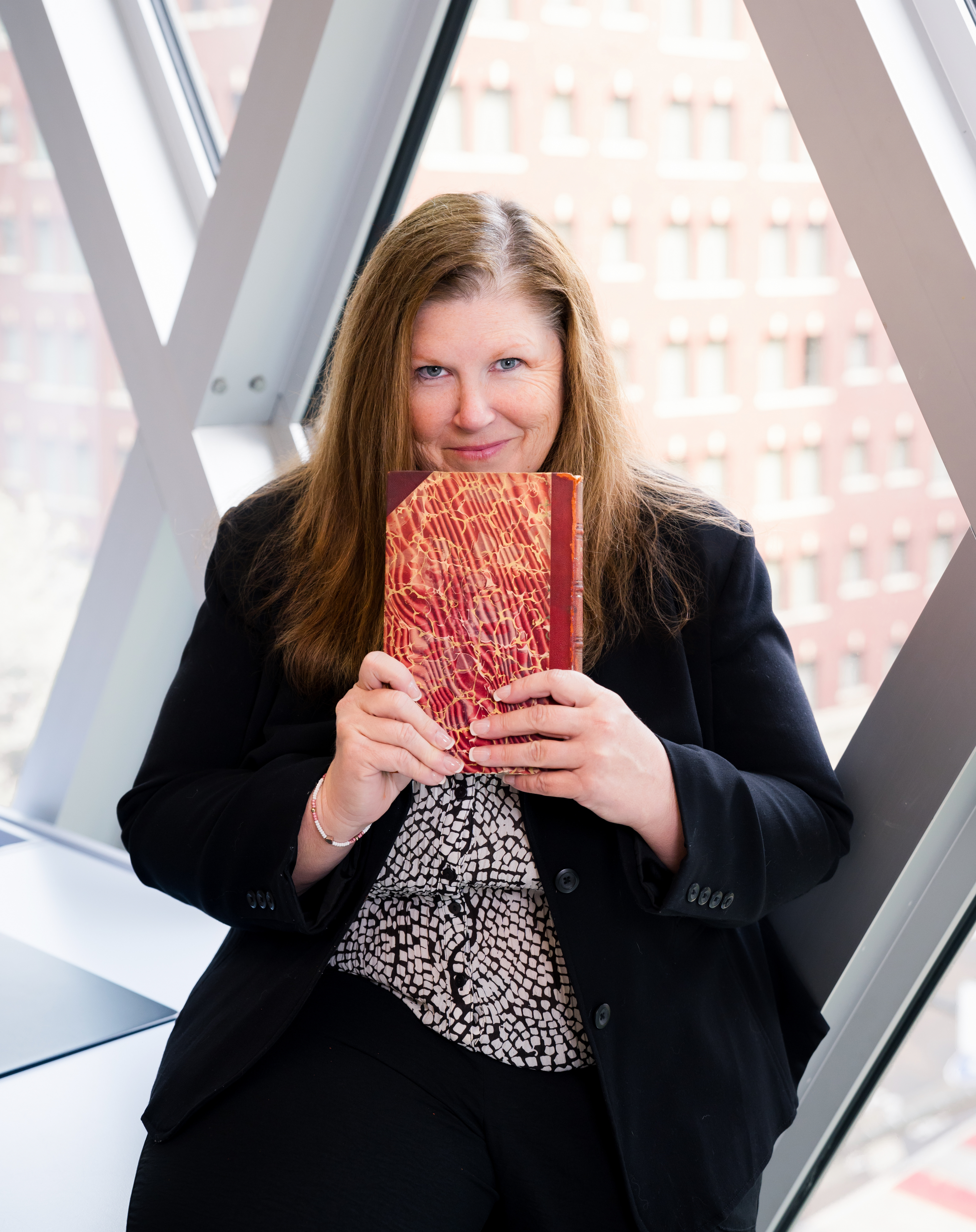 A woman dressed in a business suit is sitting in front a large window. She's holding a book with a decorative red and gold cover in front of her and peering over the top of it with a grin on her face.