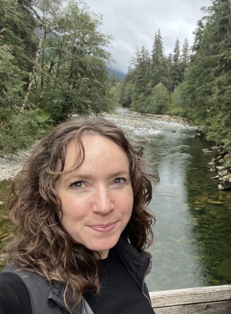 A selfie image of a woman standing in front of a river surrounded by forest on either side of its banks.