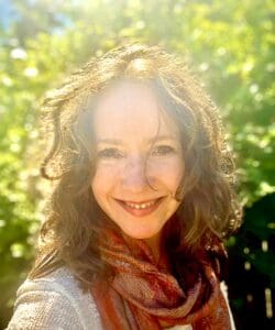 Headshot image of a woman in full color. She has a white compplexion and auburn hair. She is smiling big at the camera and standing in front of a bright green tree.